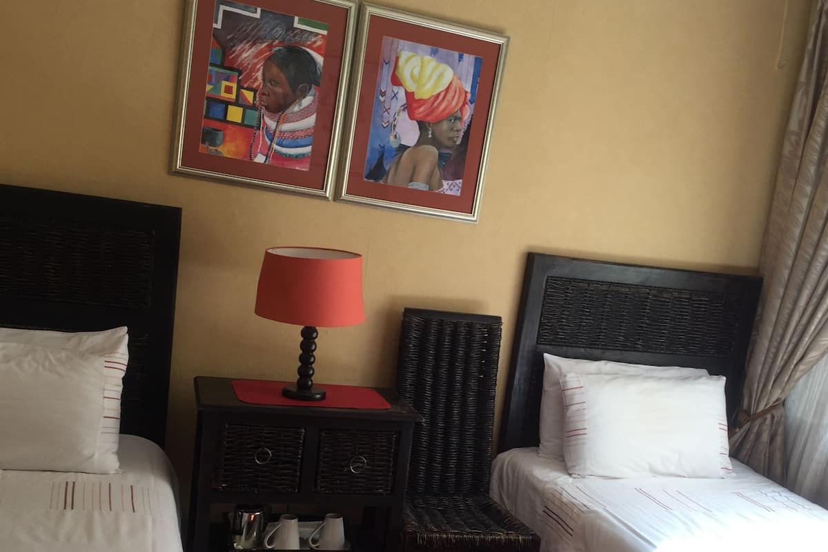 A hotel room with two beds, a nightstand with a lamp, a small table with cups and a kettle, and two framed pictures on the wall.