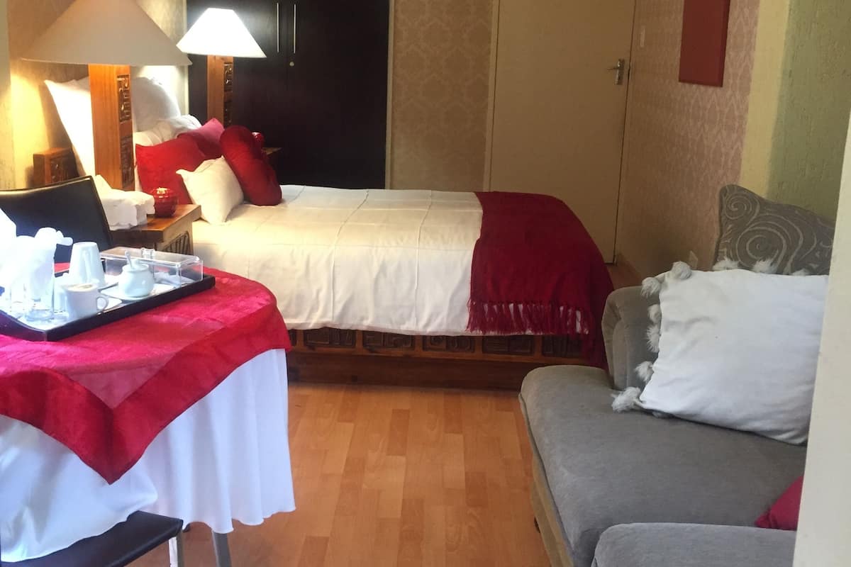 A small, cozy hotel room with a single bed, a gray sofa, a small table with a tray of breakfast items, and a lamp.
