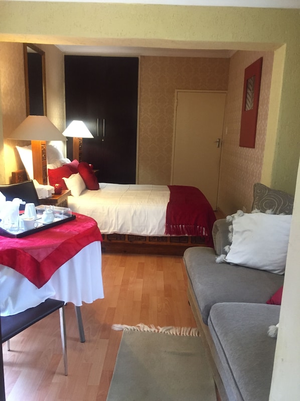 A small, cozy hotel room with a single bed, a gray sofa, a small table with a tray of breakfast items, and a lamp. - Dithakong Bed & Breakfast (Soweto)