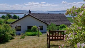 A house with a sloped roof, a chimney, and a garden with a bench. - Steading Holidays - Ardvaig (Acharacle)