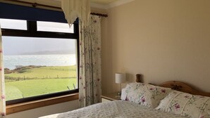 A bedroom with a bed, a nightstand, a lamp, and a view of the sea through a window with curtains. - Steading Holidays - Ardvaig (Acharacle)