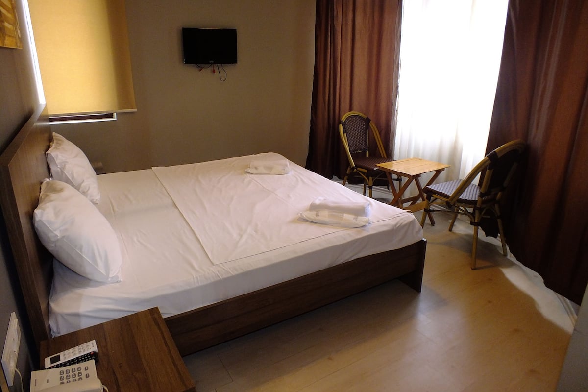 A hotel room with a single bed, a small table, two chairs, a TV mounted on the wall, and a window with curtains.