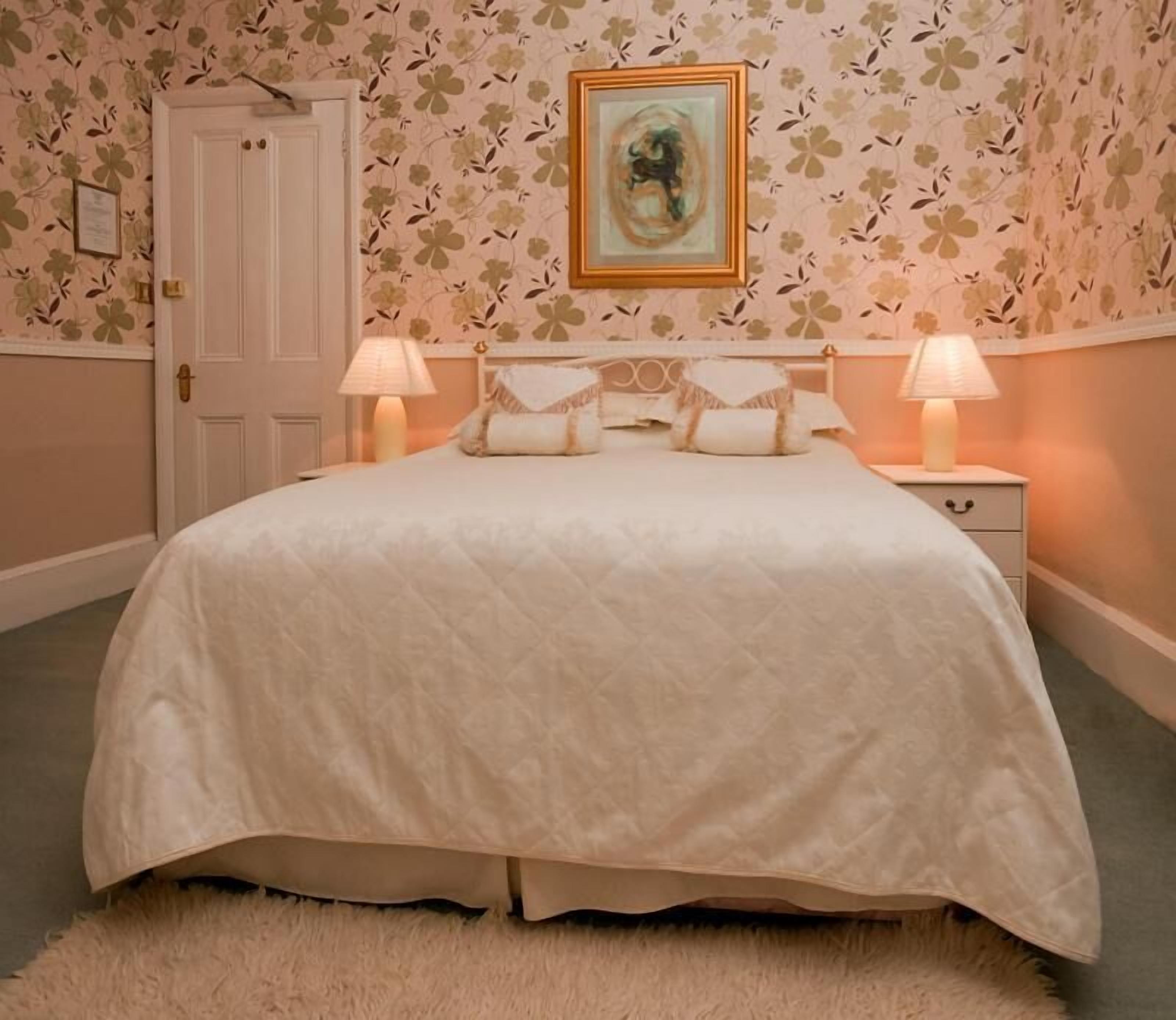 A bedroom with a bed, two bedside lamps, a door, and a floral wallpaper.
