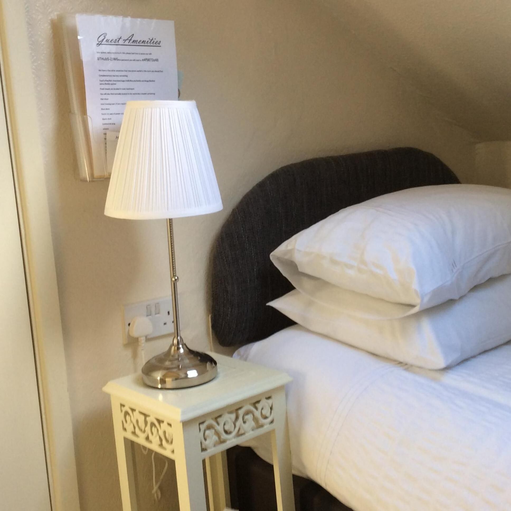 Una cama bien hecha con sábanas y almohadas blancas, una lámpara de noche sobre una mesita blanca pequeña y un letrero en la pared que dice 'Guest Amenities'.