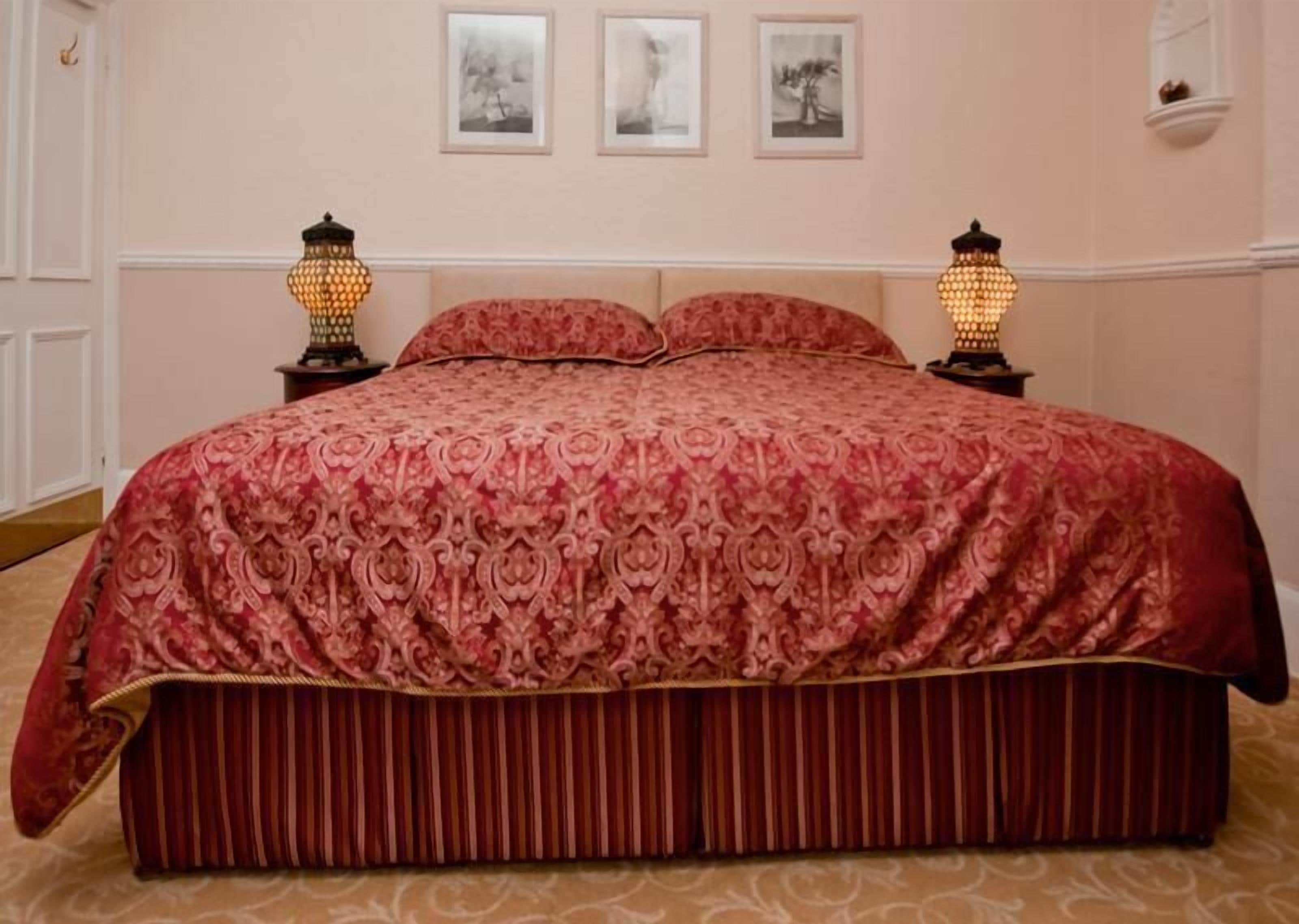 Een groot bed met een rode, gemêleerde spreideken en passende kussens, aan weerszijden verlicht door twee wandlampen.