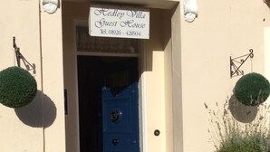 Ein beiges Gebäude mit einer blauen Tür, einem Schild mit der Aufschrift "Hedley Villa Guest House" und zwei in Töpfen stehenden Formschnitten.