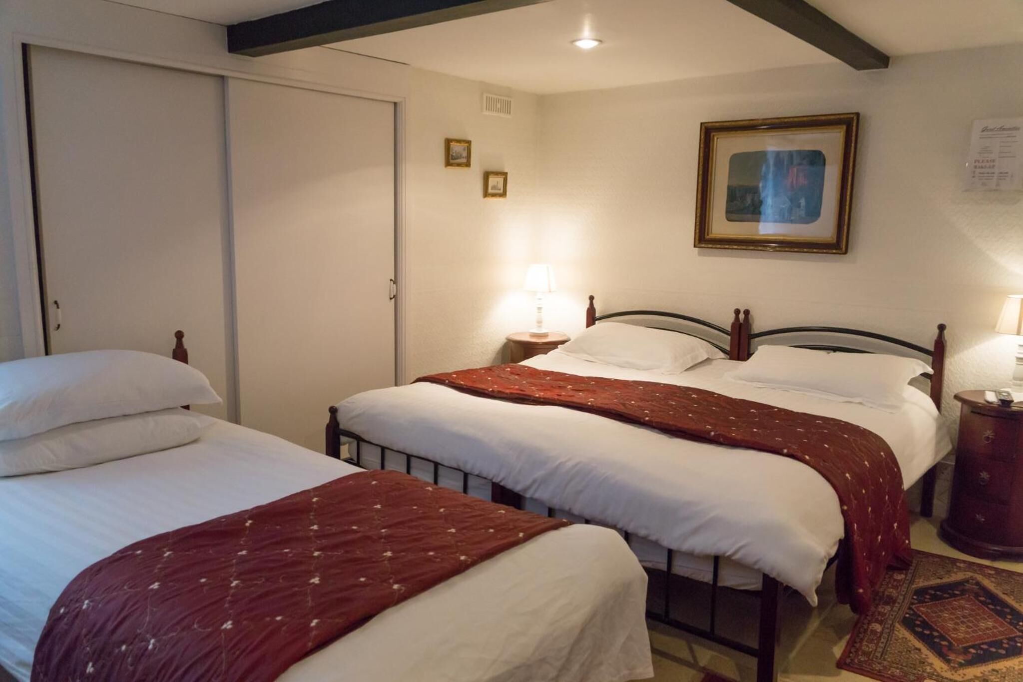 A hotel room with two single beds, a wooden headboard, a nightstand with a lamp, a framed picture on the wall, and a small rug on the floor.