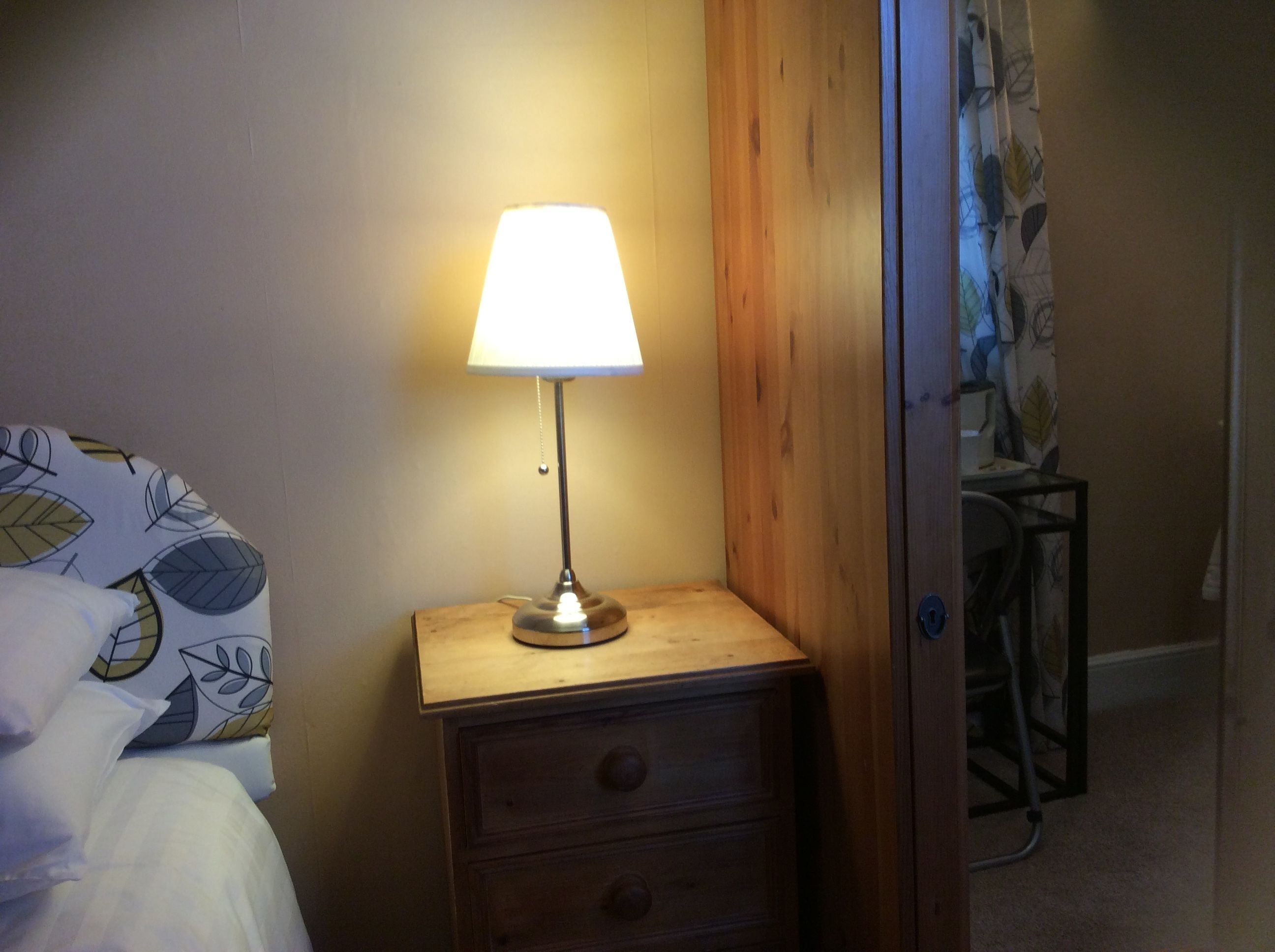 Ett litet bord vid sängen med en lampa, ett träbord som står bredvid sängen, en stol och en säng med mönstrat sänglinne.