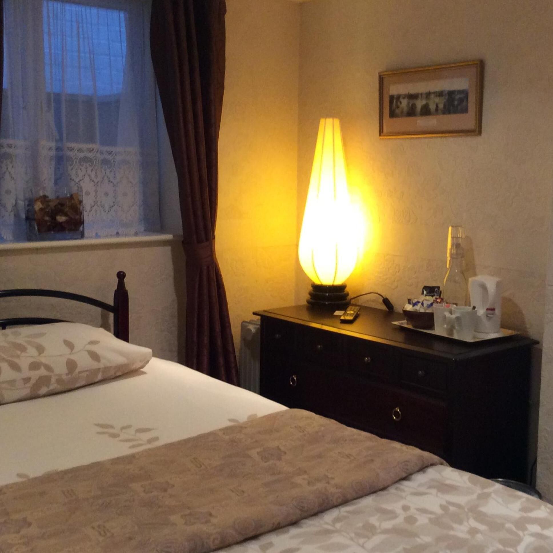 A hotel room with a bed, a bedside table with a lamp, a framed picture on the wall, and a window with curtains.