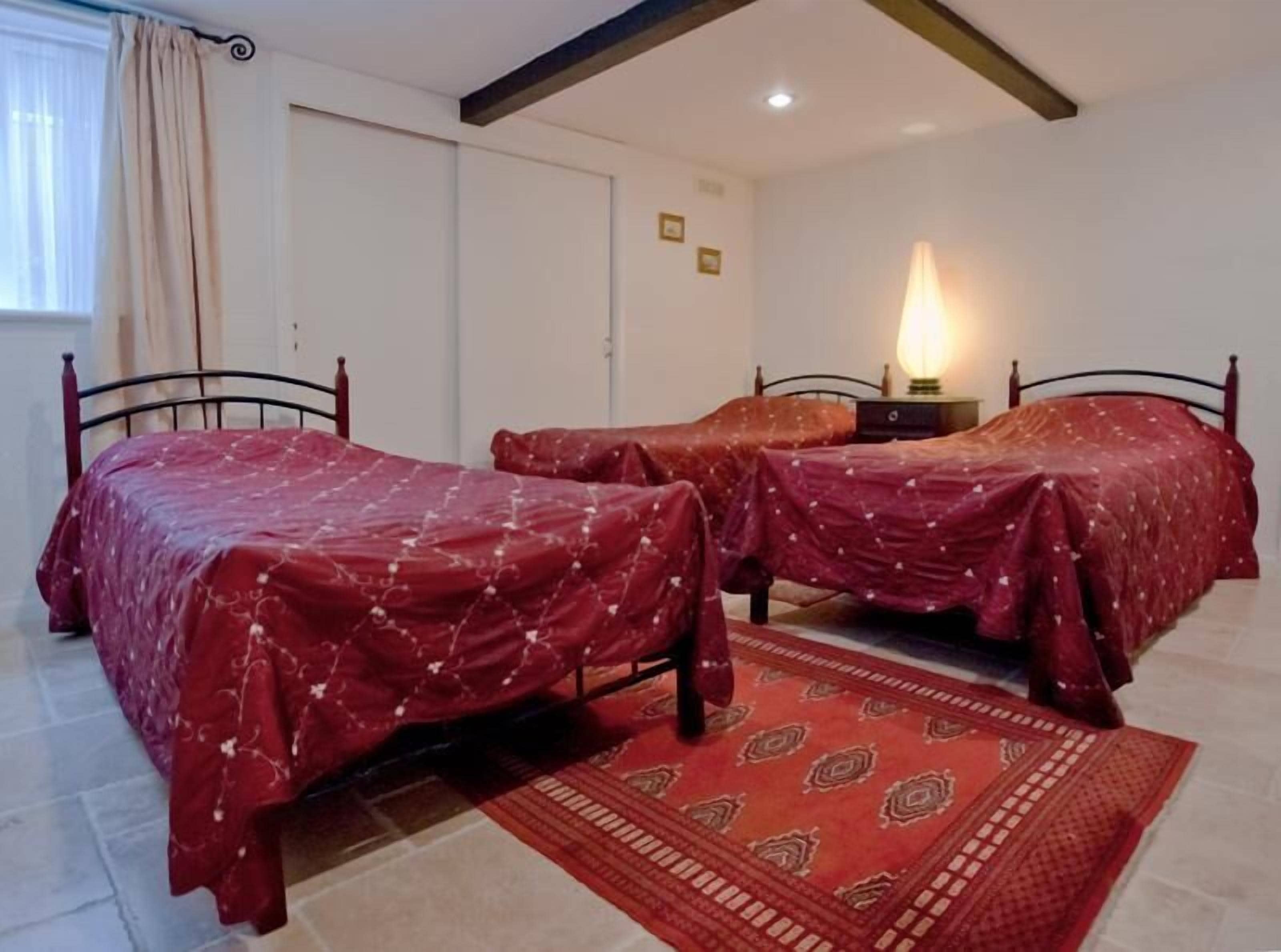 Una habitación con dos camas, un tapete estampado, una mesita de noche con lámpara y una ventana con cortinas.