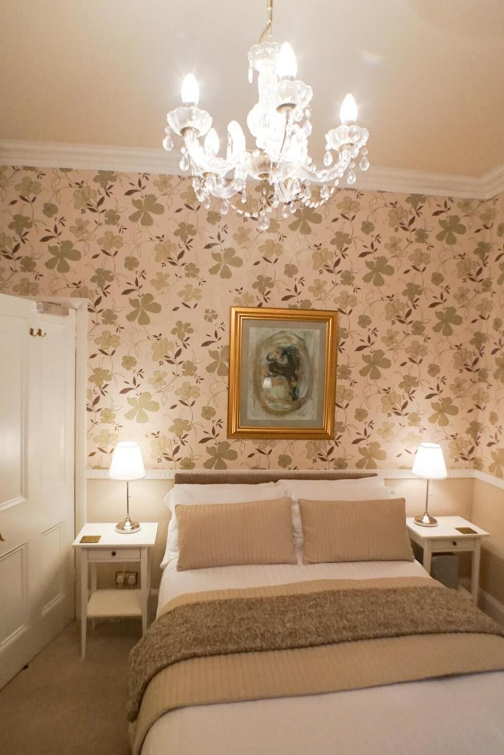 Un dormitorio con una cama, dos mesitas de noche con lámparas, una araña, papel tapiz floral y un cuadro en la pared.