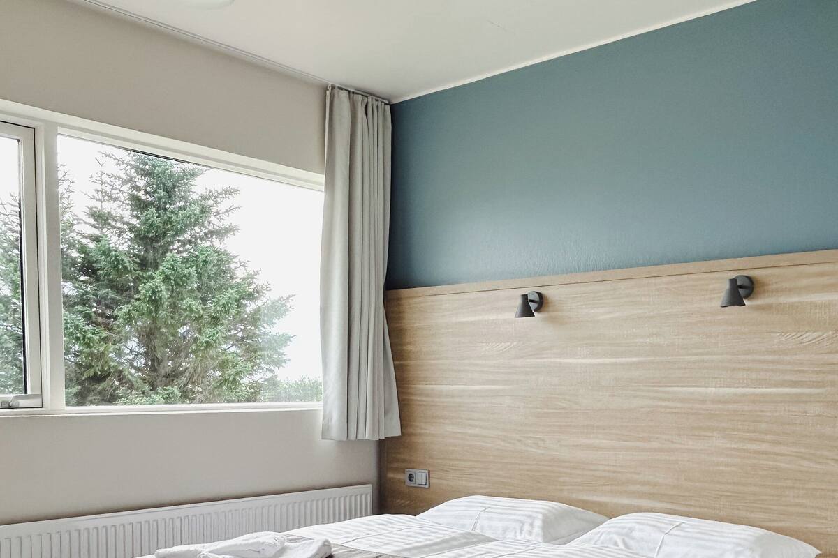 A neatly made bed with white linens and a brown throw blanket, a window with curtains, and a light blue wall.