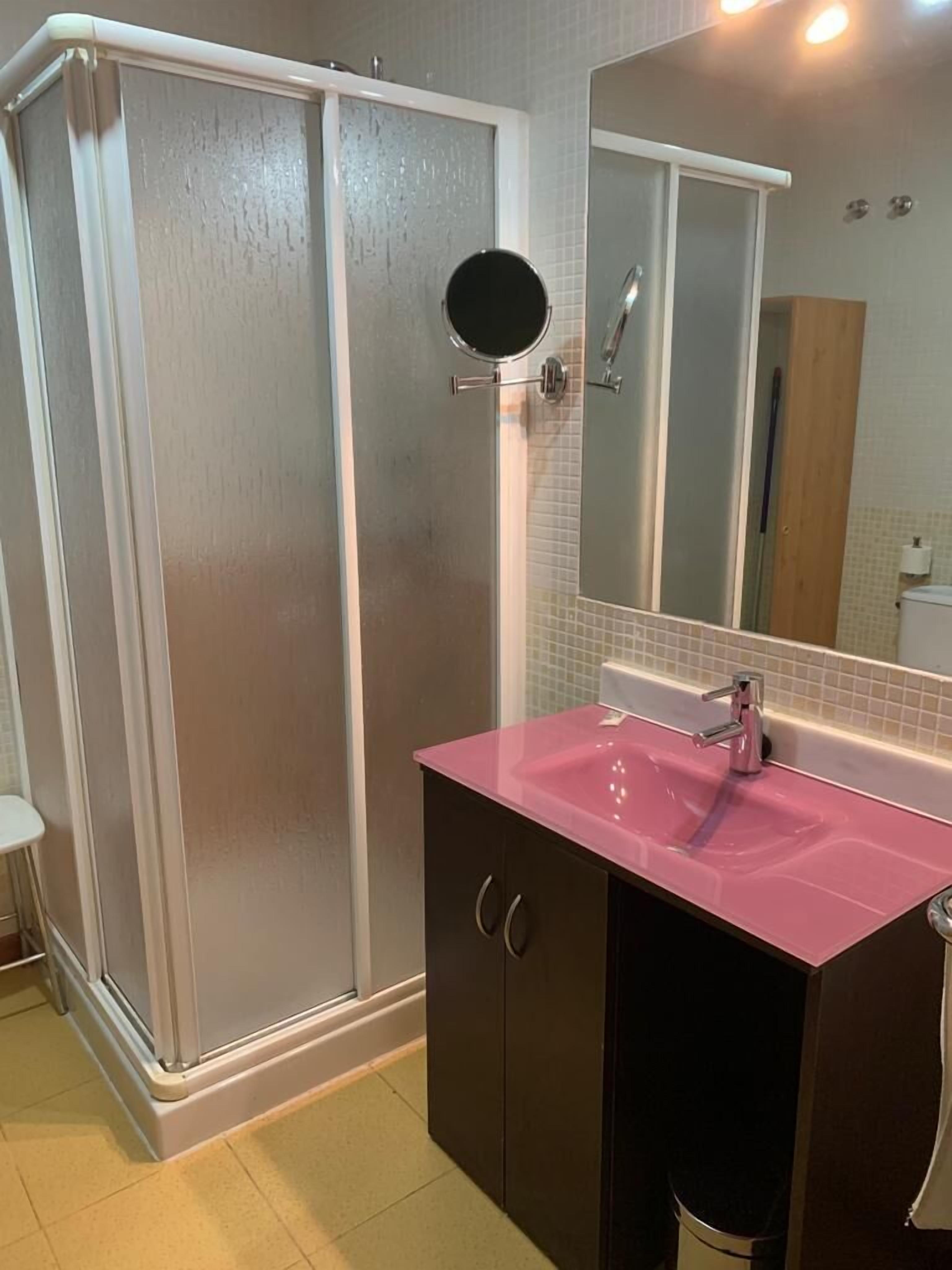 Une salle de bain avec une douche à l’italienne, un lavabo rose et un miroir au-dessus.