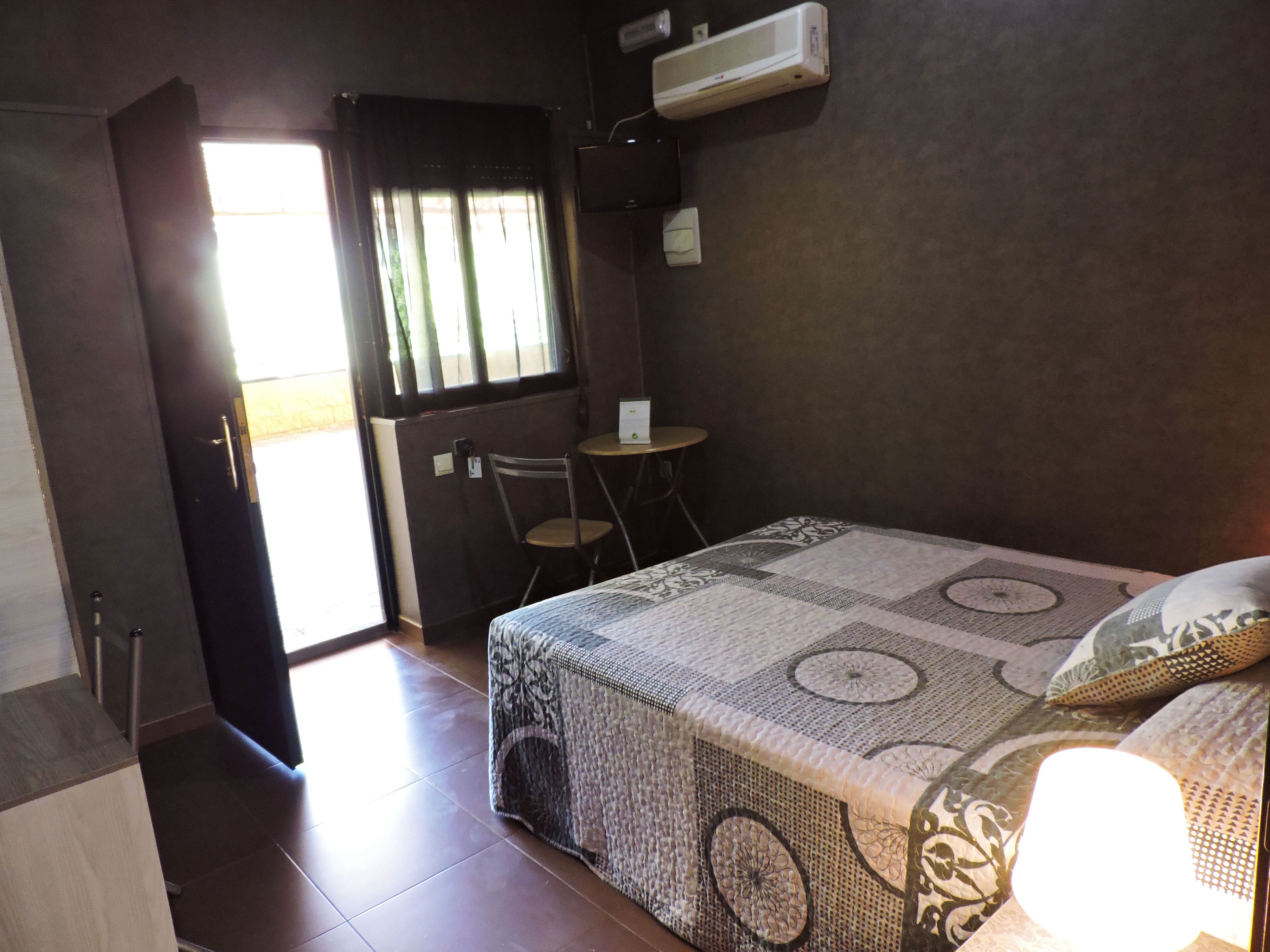 Una habitación pequeña y con poca luz, con una cama, una silla, una mesita y un equipo de aire acondicionado.