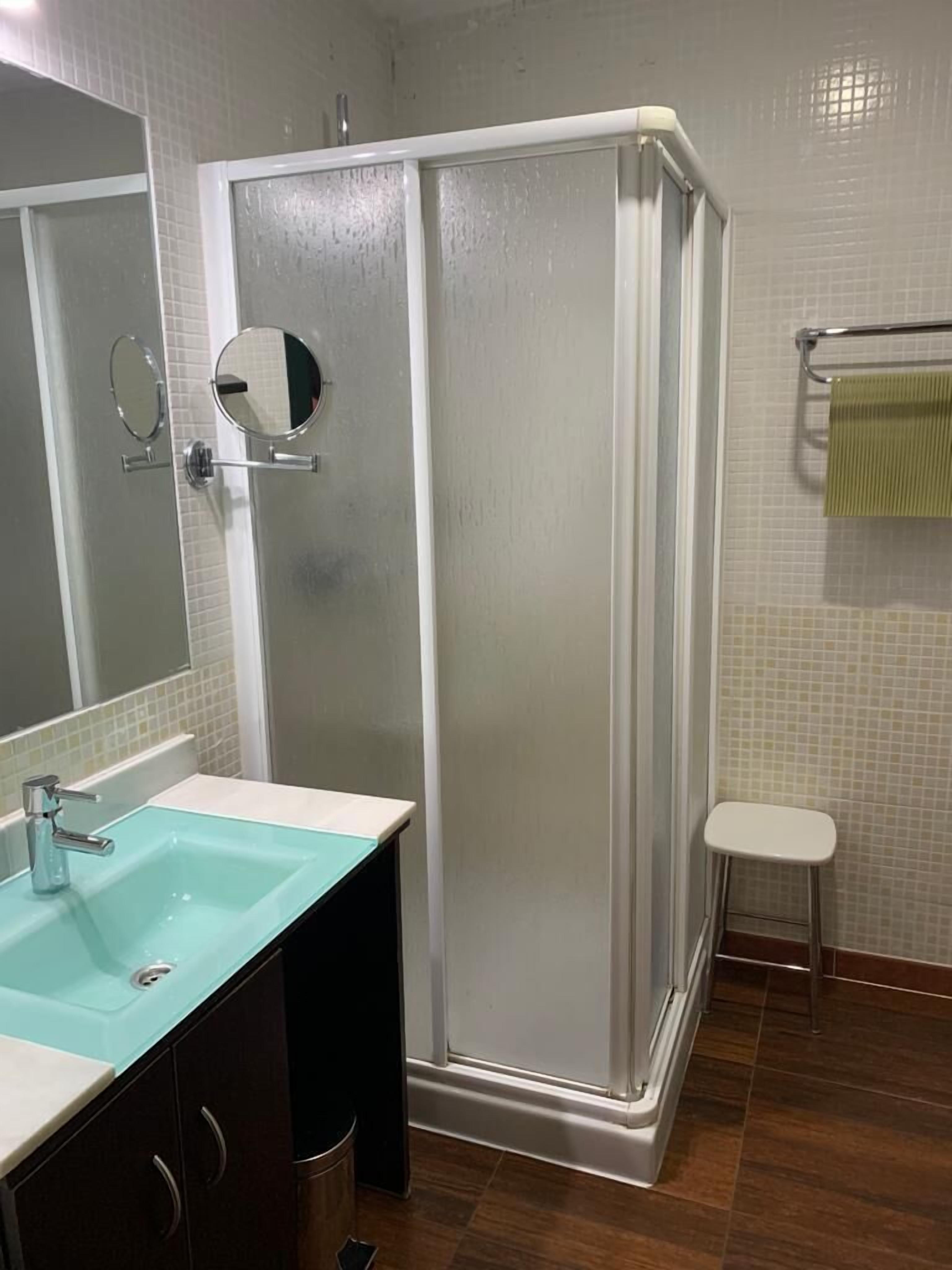 Een badkamer met een douchecabine van glas, een wastafel met spiegel en een klein krukje.
