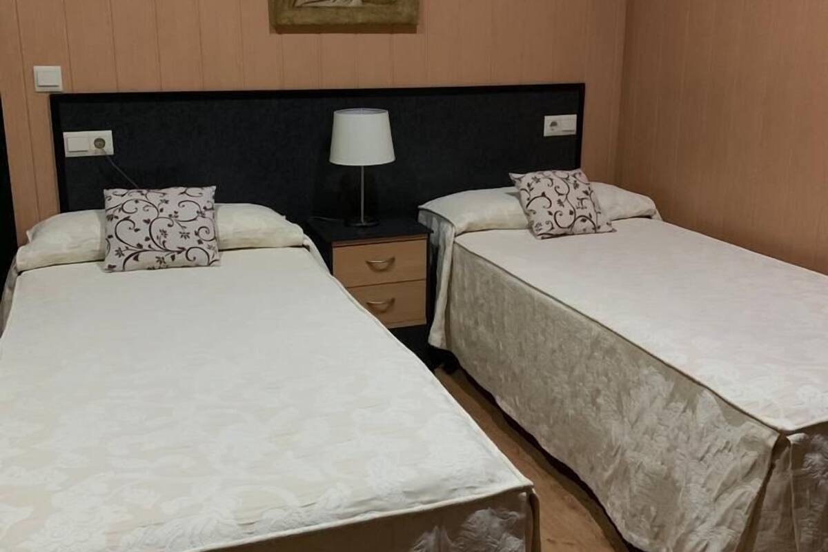 A hotel room with two single beds, a nightstand with a lamp, a wall-mounted painting, and wooden paneling.