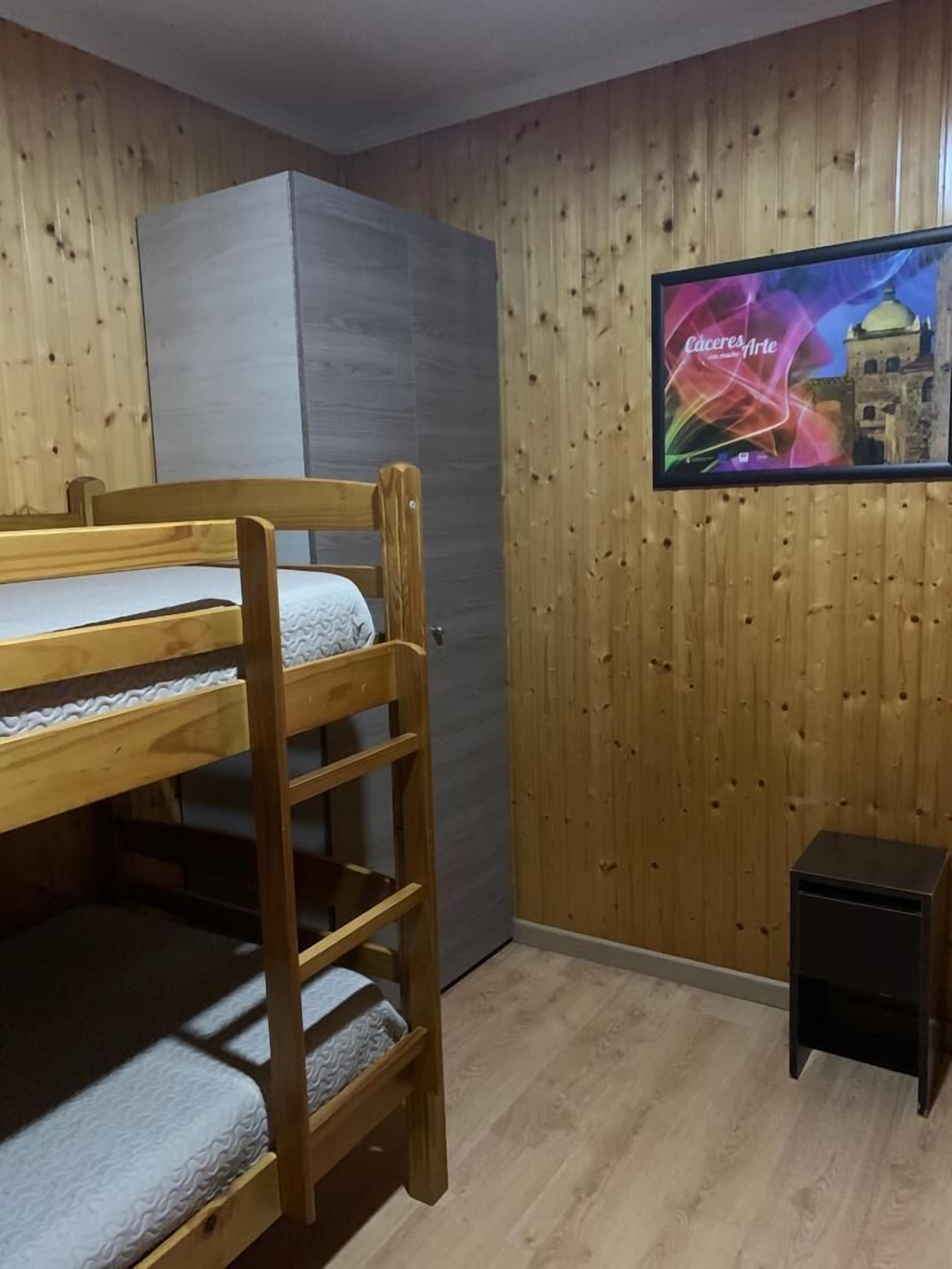 Una habitación con una cama nido, un armario gris y un televisor mostrando 'Cáceres Arte'.