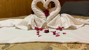 Una cama con cubrecama blanco y un arreglo de toallas con forma de corazón y pétalos de rosa.