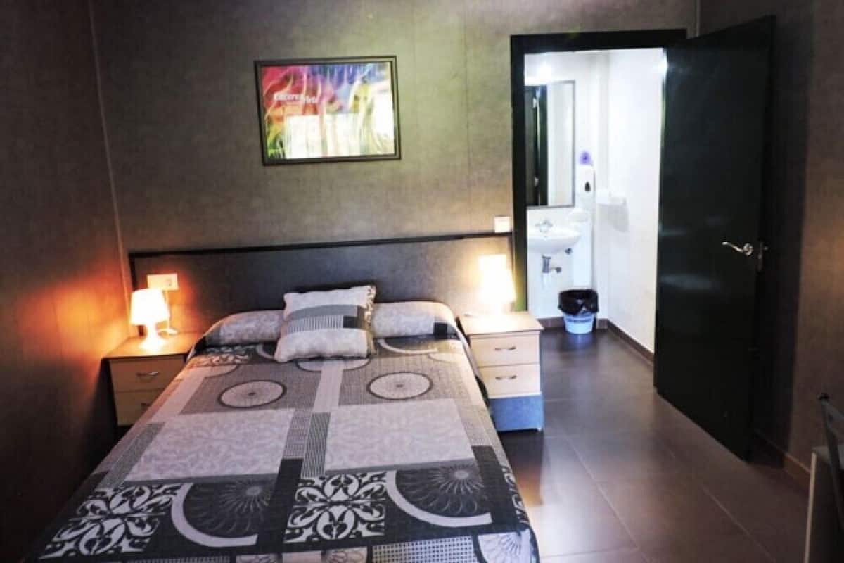 A bedroom with a bed, bedside tables, a nightstand, a wall-mounted TV, and a door leading to a bathroom.