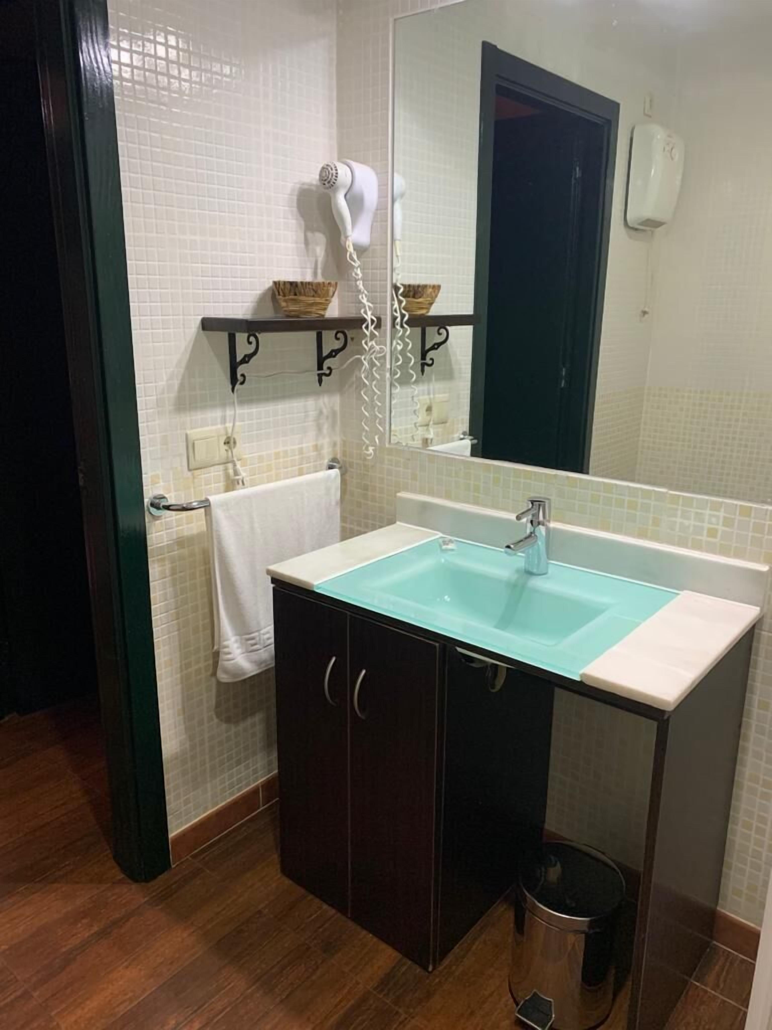 Un baño con lavamanos de vidrio, muebles oscuros, una toalla blanca, un secador de pelo y un bote de basura.