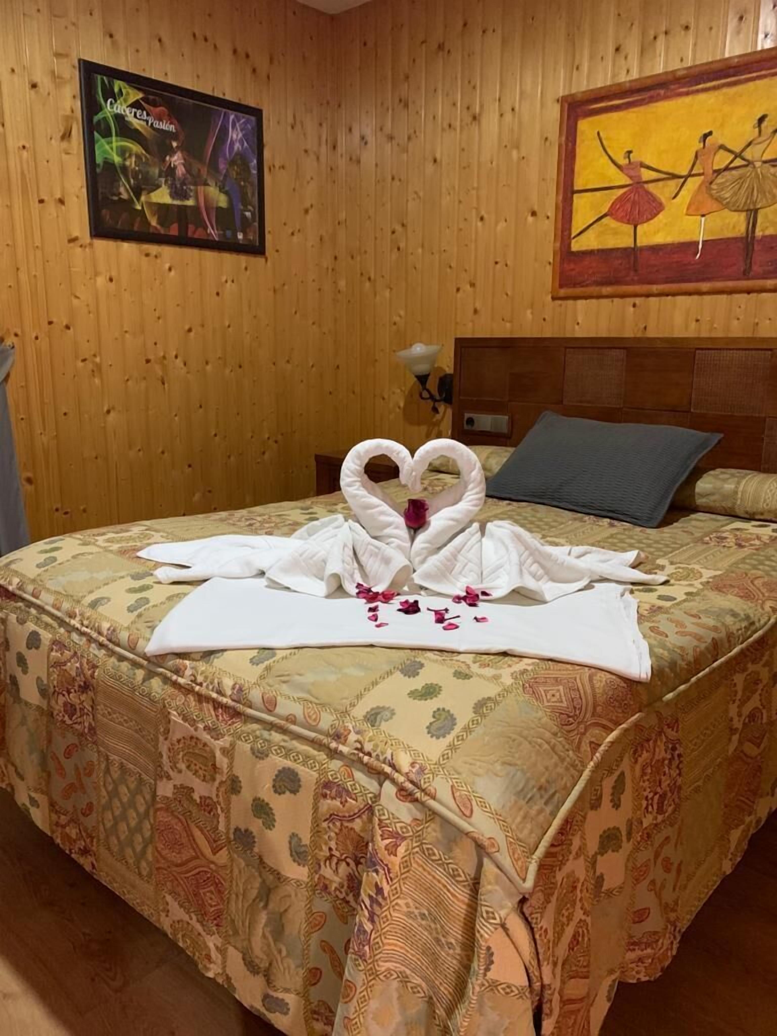 Hotellihuone, jossa on puupanelointi yhdellä seinällä, kukallinen päiväpeite sängyllä, sydämenmuotoinen pyyheasetelma ja tanssijoiden kuva.