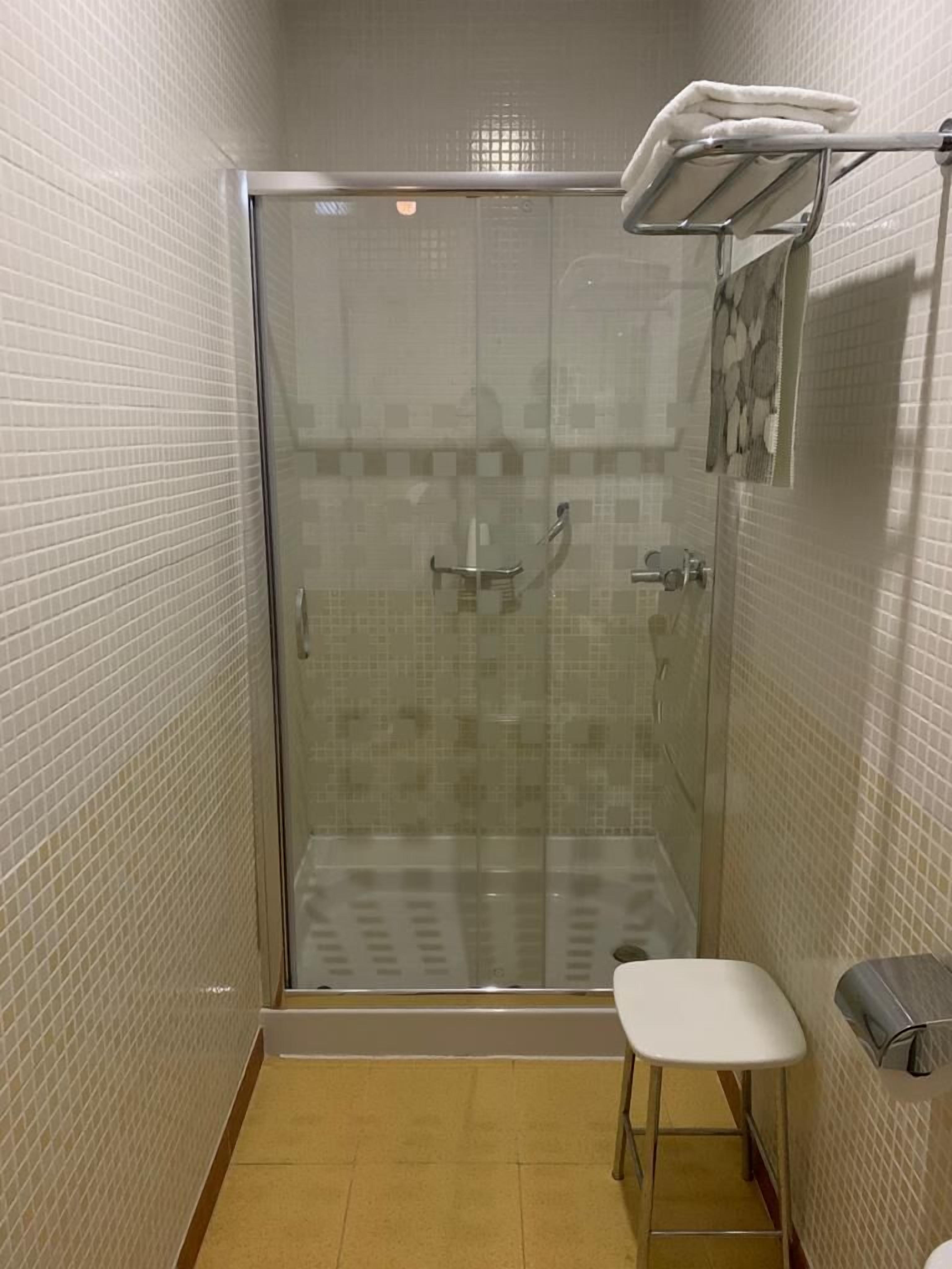Un área de ducha pequeña y cerrada con puerta de vidrio, un banco blanco y un perchero para toallas.
