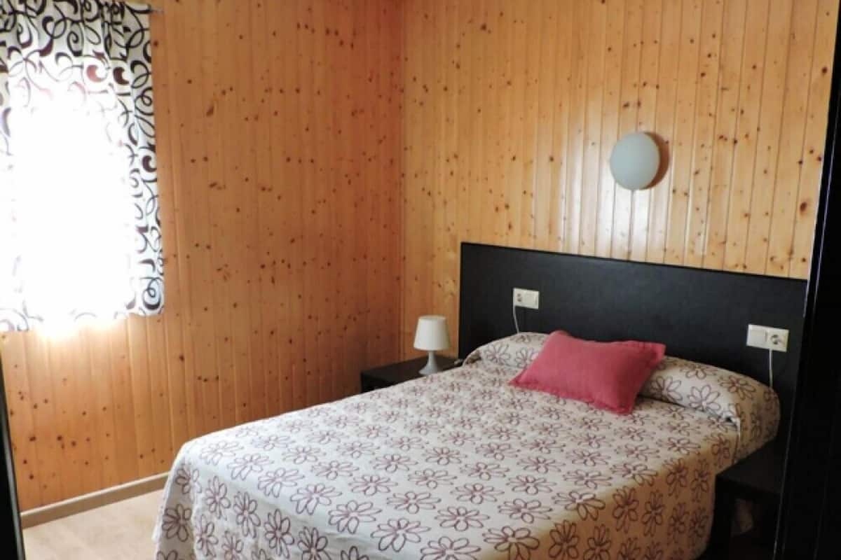 A bedroom with wooden panel walls, a bed with a floral bedspread, a round wall clock, and a nightstand with a lamp.