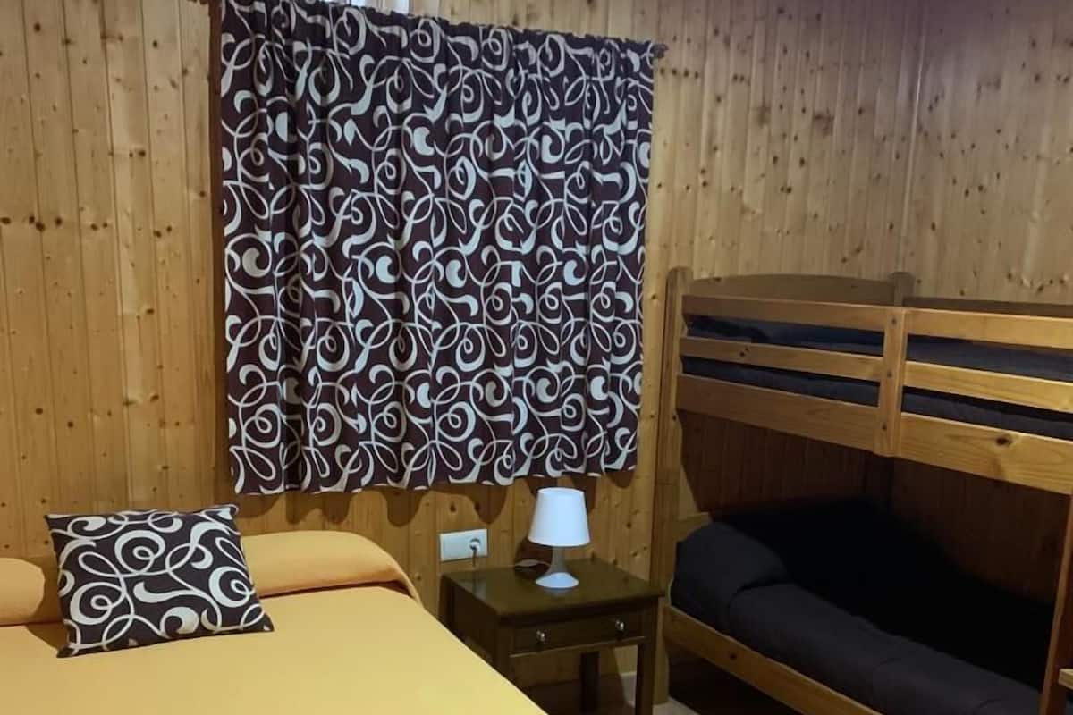 A room with a bunk bed, a single bed with a patterned pillow, a bedside table with a lamp, and wooden walls.