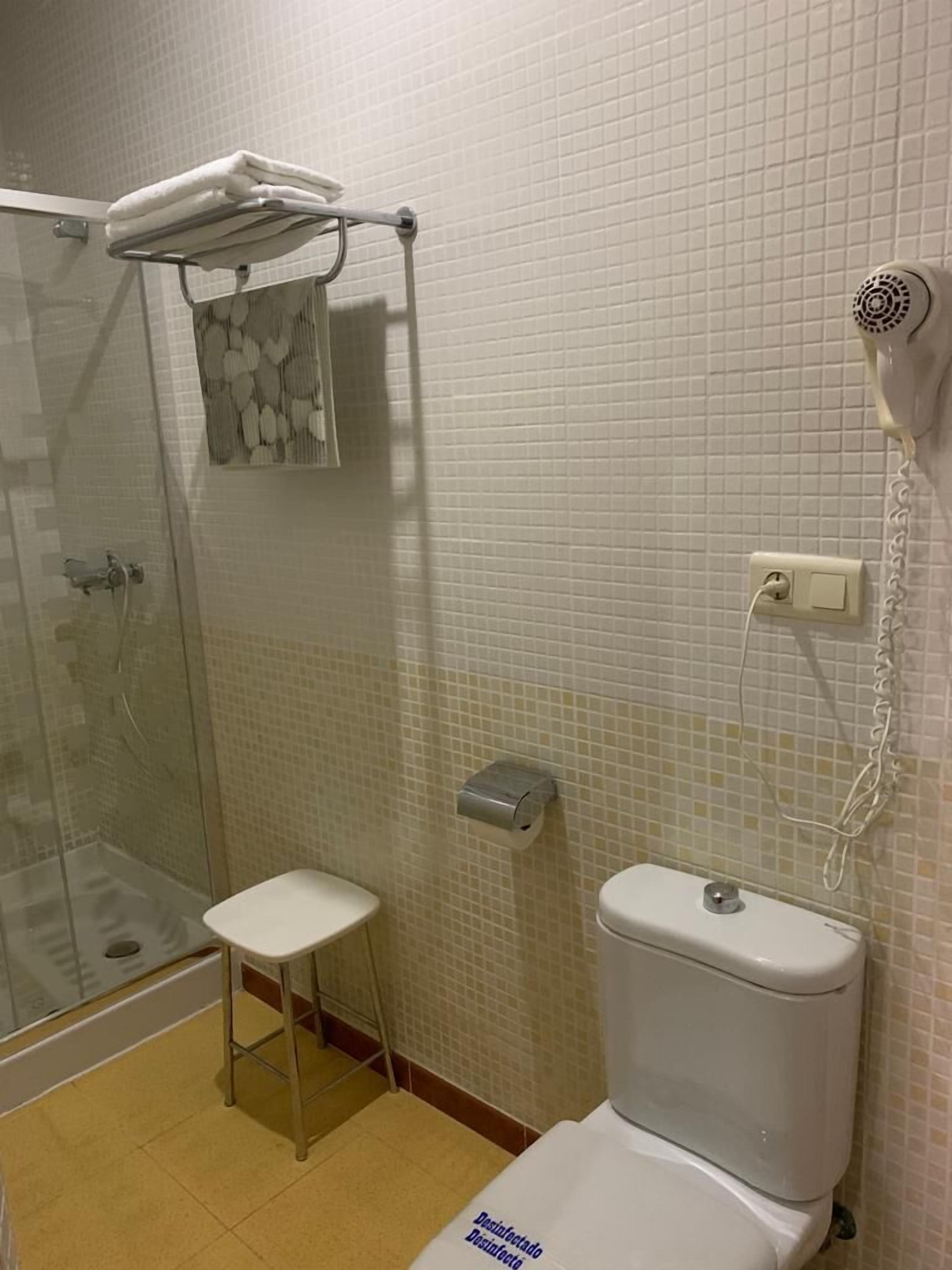 Un baño con cabina de ducha de vidrio, inodoro, un taburete pequeño y un perchero para toallas.