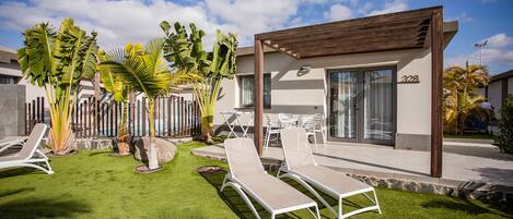 Uma área de estar moderna ao ar livre, com espreguiçadeiras brancas e uma pérgola de madeira, rodeada por plantas tropicais.