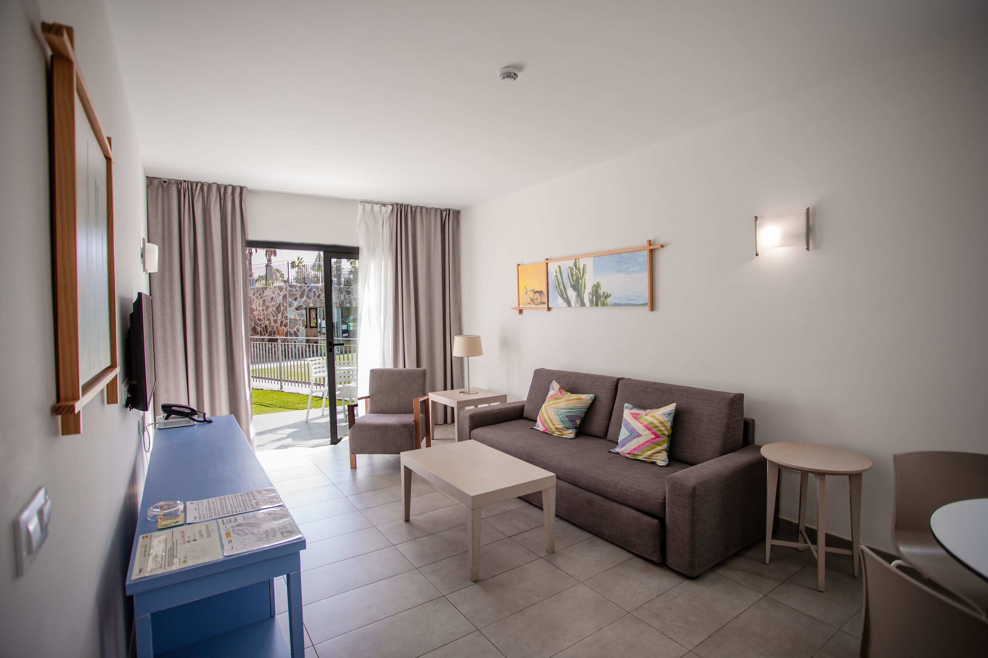 Ein modernes Wohnzimmer mit einer Couch, einem Couchtisch und Sesseln. Es gibt einen Schreibtisch mit einer Lampe und einem Fernseher. Ein Gemälde hängt an der Wand, und ein Fenster mit Vorhängen ist sichtbar.
