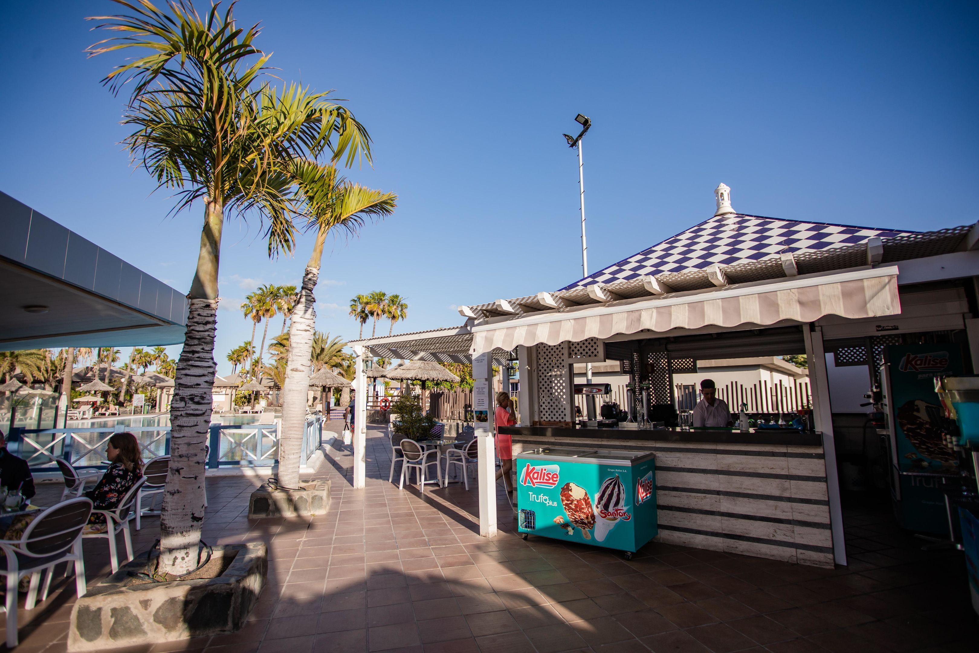 An outdoor seating area with palm trees, a kiosk, and a view of a building with a checkered roof.