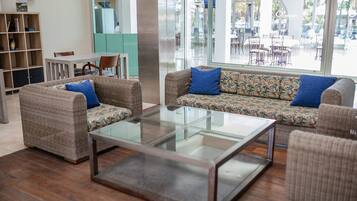 Una sala de estar moderna con una mesa de centro de vidrio, dos sofás con cojines estampados, una estantería de madera y vistas a un área de comedor exterior a través de ventanas amplias.