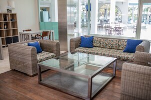 Una sala de estar moderna con una mesa de centro de vidrio, dos sofás con cojines estampados, una estantería de madera y vistas a un área de comedor exterior a través de ventanas amplias.