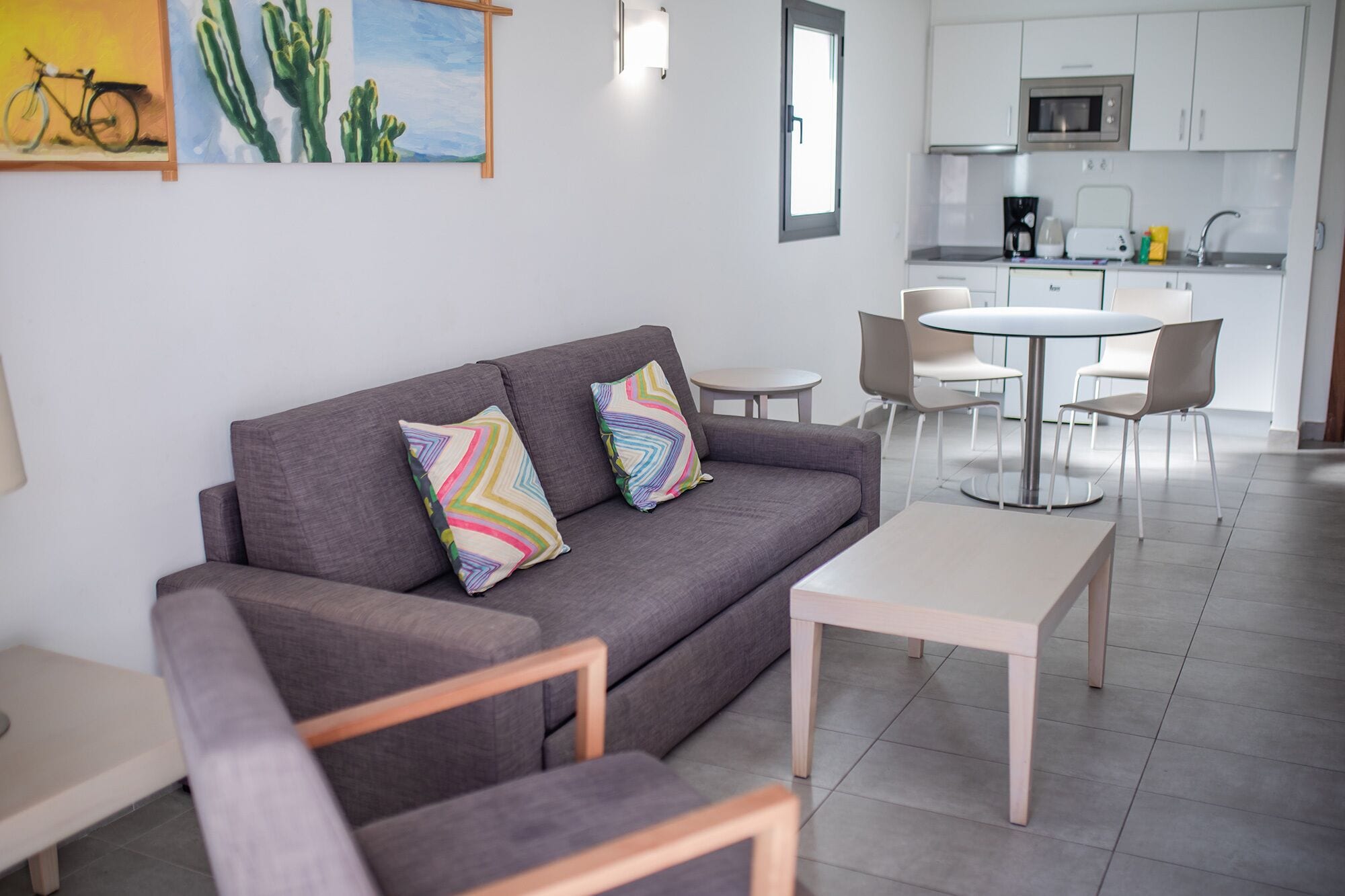 Een moderne woonkamer met een grijze bank, twee kleurrijke kussens, een klein witte tafel en een eethoek met een ronde tafel en stoelen.