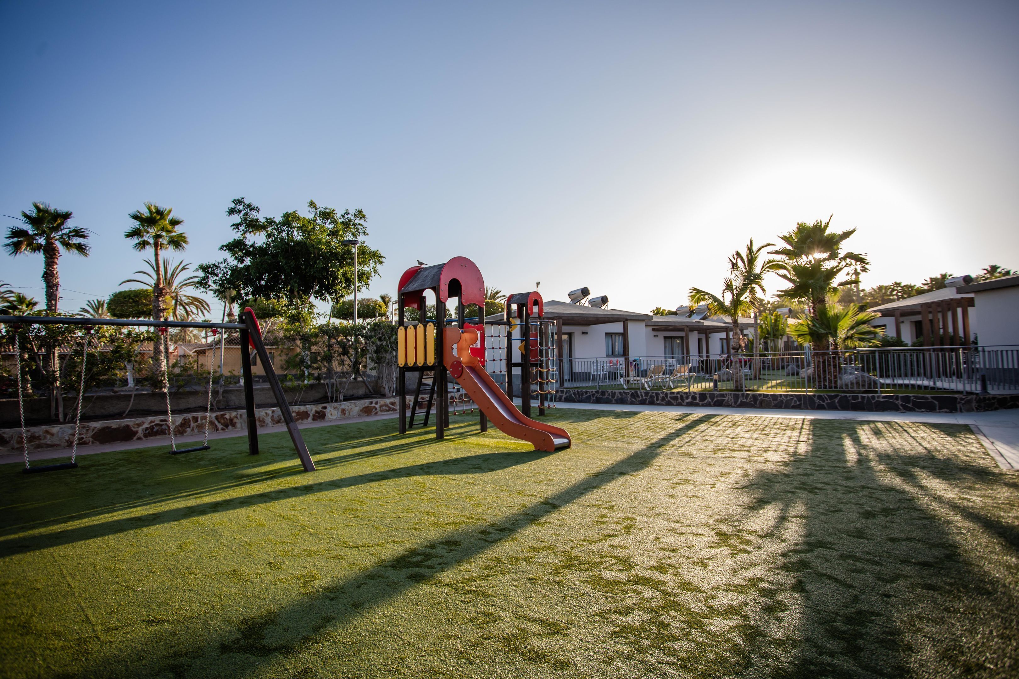 Un parque infantil con columpios y un tobogán, rodeado de palmeras y casas.