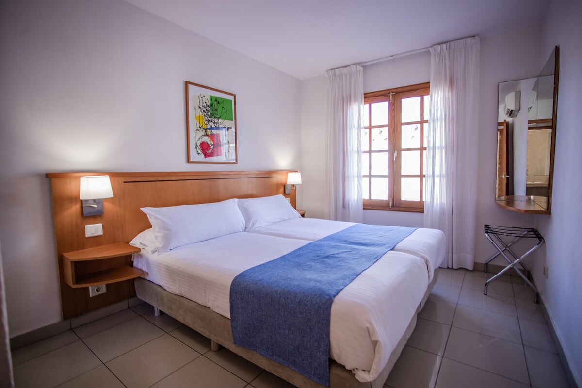 Habitación de hotel con cama doble, dos lámparas de noche, cabecera de madera, ventana con cortinas, espejo en la pared y un cuadro enmarcado.