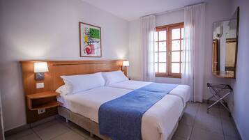 A hotel room with a double bed, two bedside lamps, a wooden headboard, a window with curtains, a wall-mounted mirror, and a framed picture.