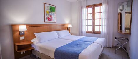 A hotel room with a double bed, two bedside lamps, a wooden headboard, a window with curtains, a wall-mounted mirror, and a framed picture.