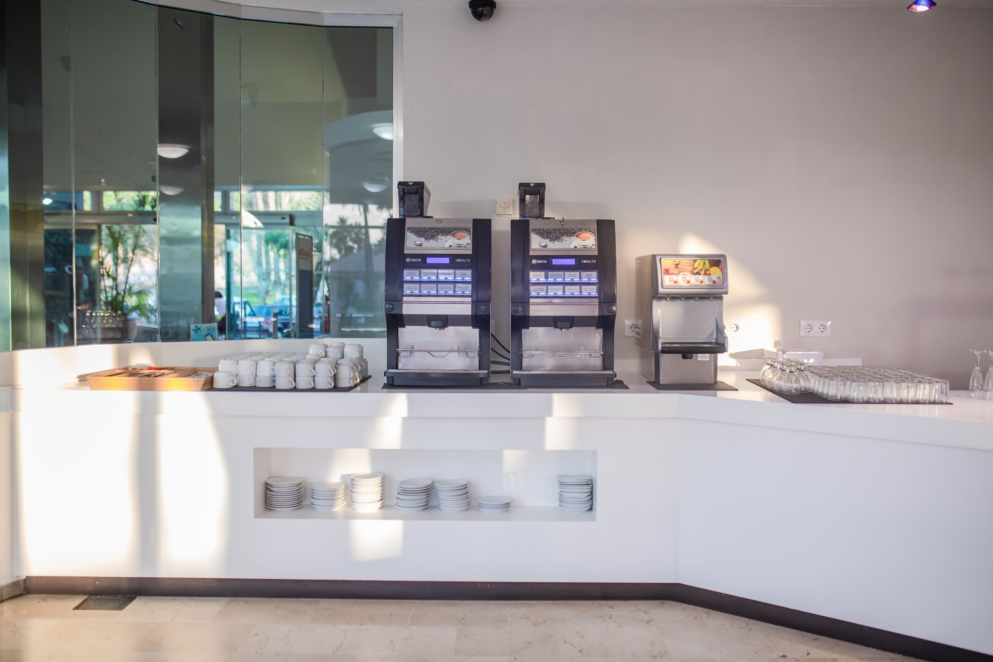 A breakfast bar with coffee machines, a fruit display, and neatly stacked plates and cups.