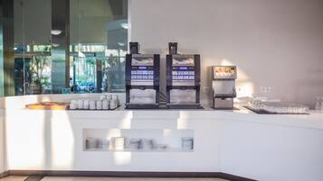 A breakfast bar with coffee machines, a fruit display, and neatly stacked plates and cups.