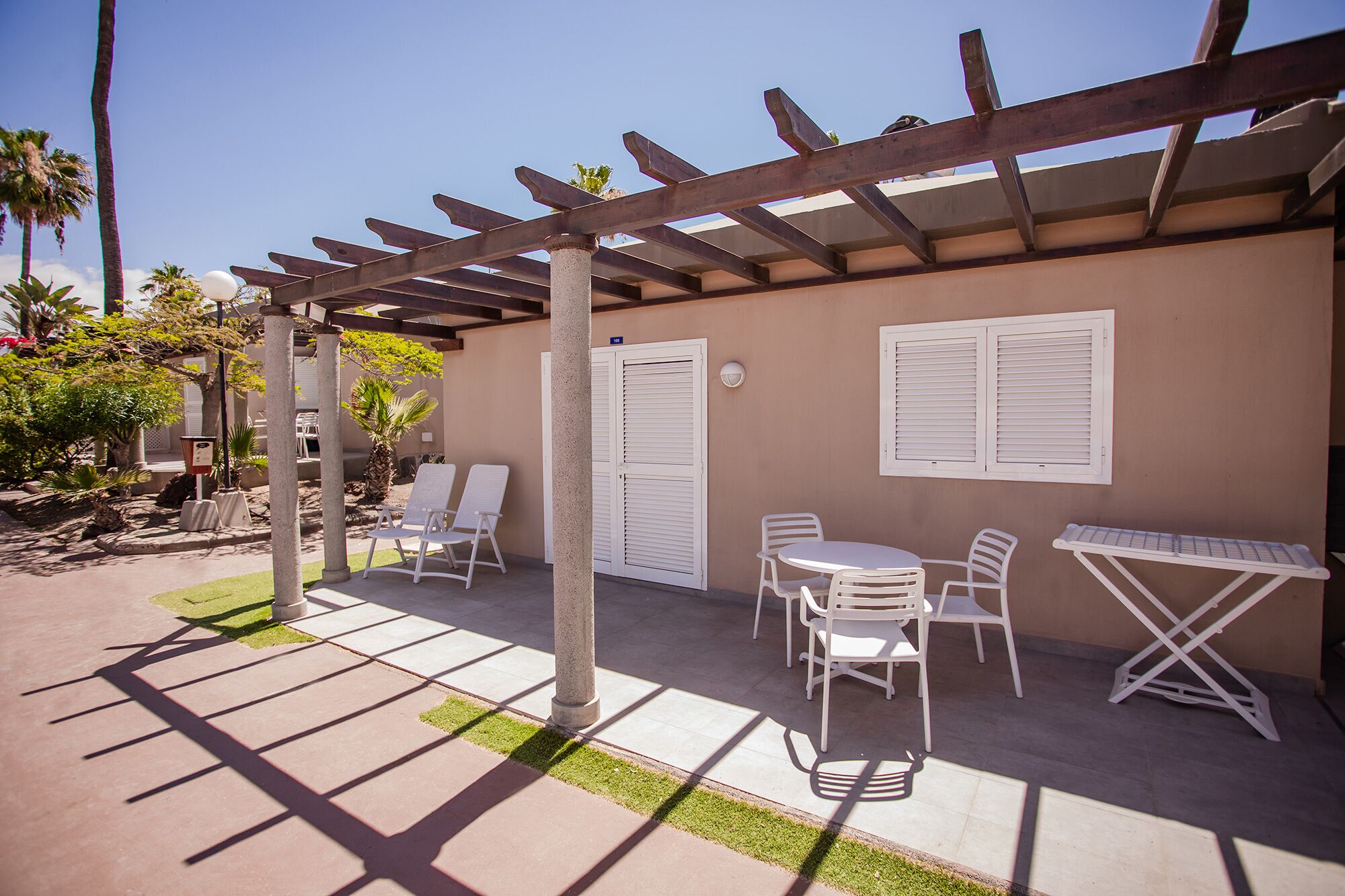 Un patio exterior con pérgola, sillas blancas, una mesa y un pequeño jardín con palmeras.