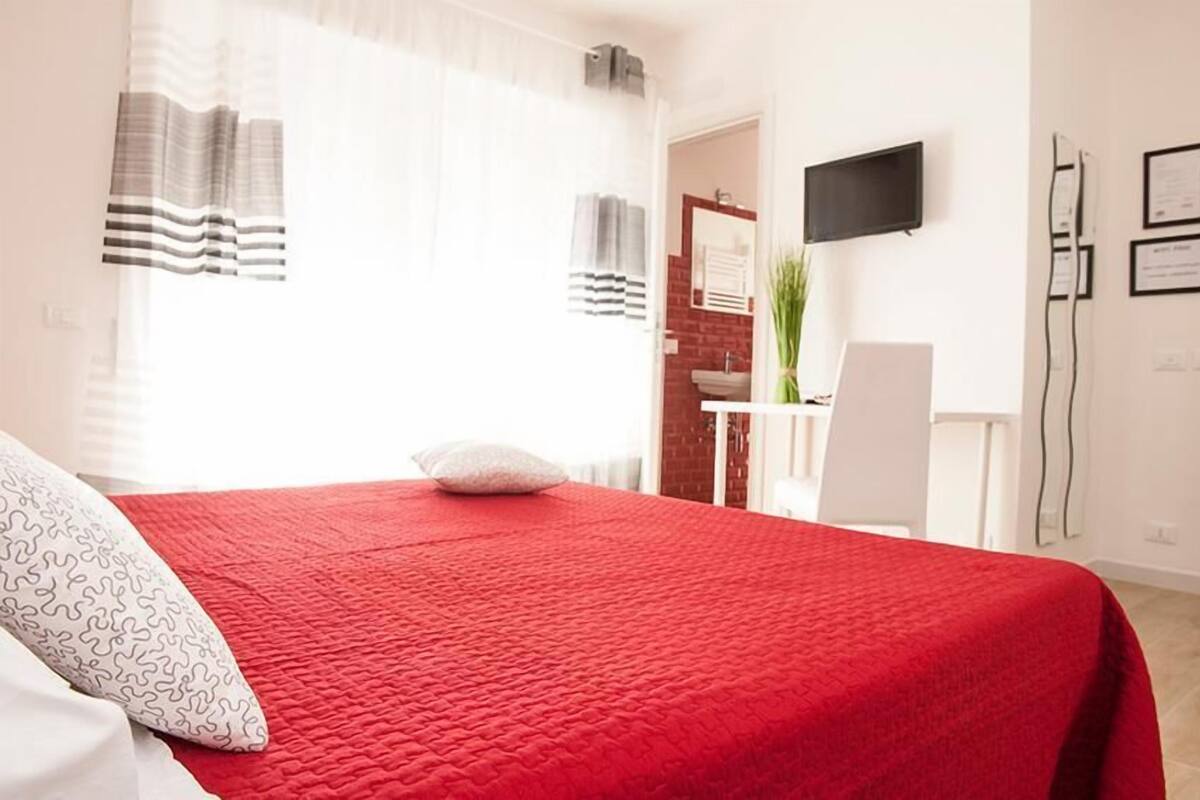 Quarto de hotel com colcha vermelha, travesseiro branco, cortina listrada, escrivaninha com cadeira, televisão na parede e uma planta.