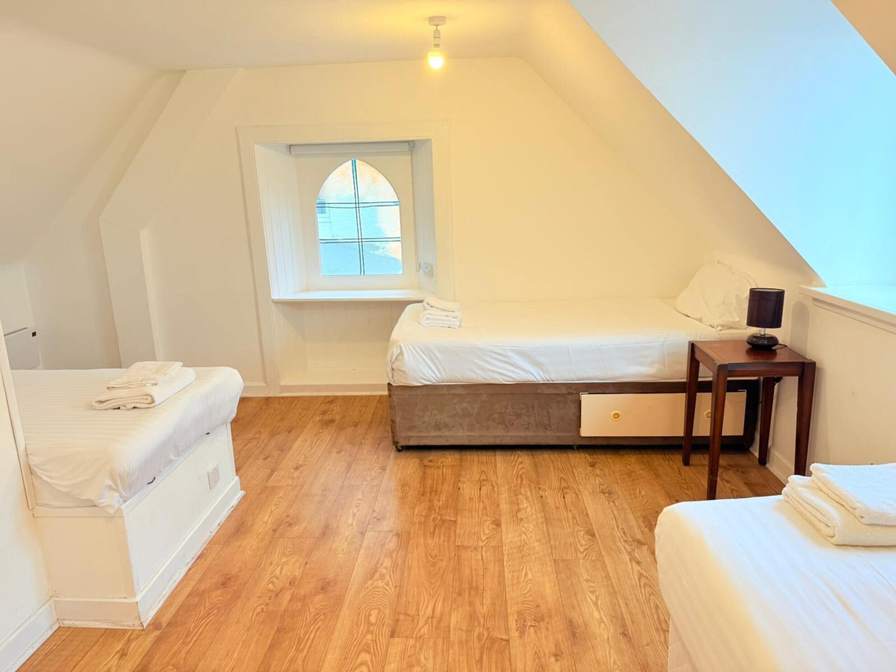 A small, well-lit room with a sloped ceiling, a single bed with a wooden headboard, a small table with a lamp, and a window with a view of the outside.