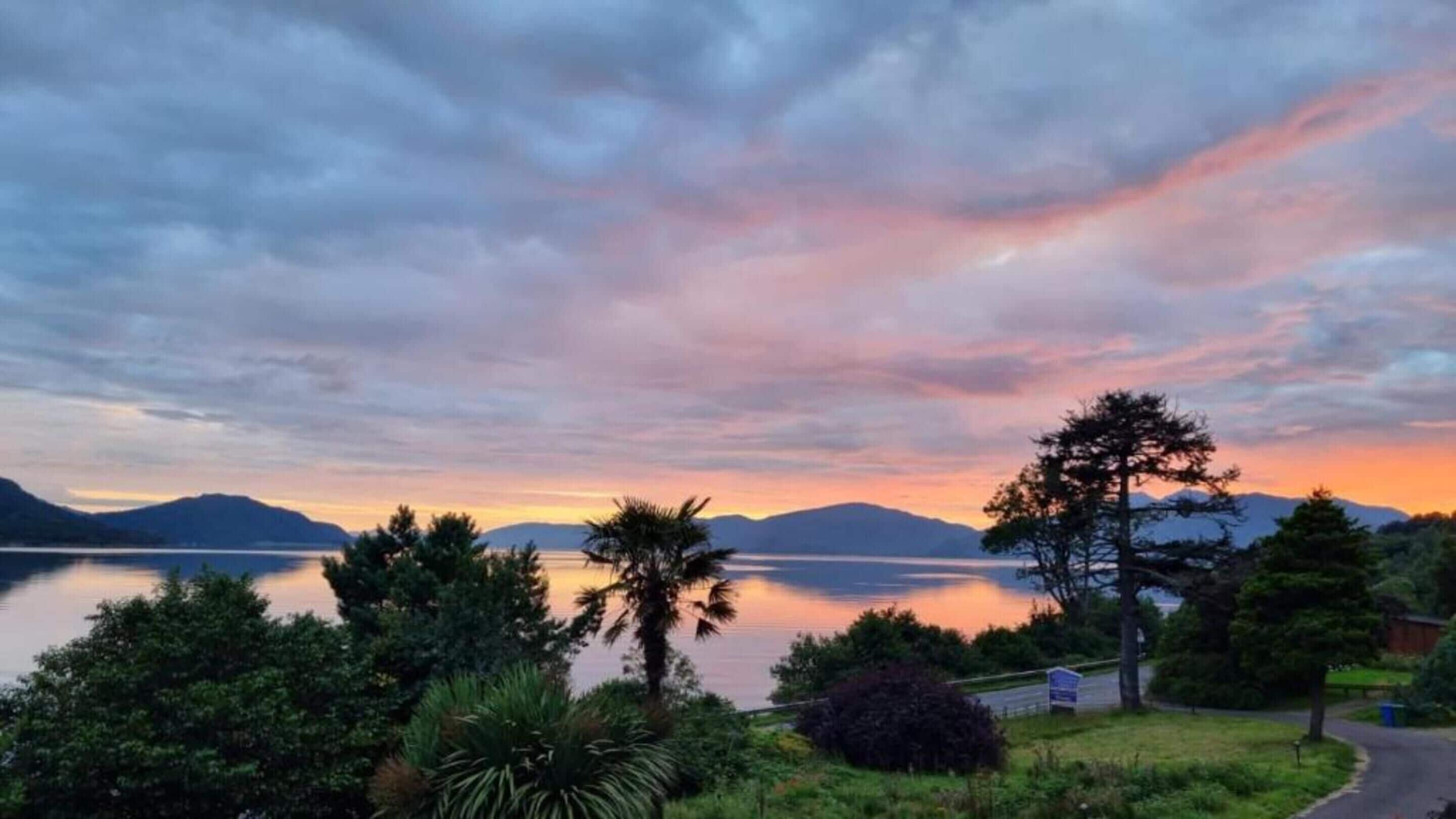 A serene lakeside view with mountains in the background, lush greenery, and a clear sky.
