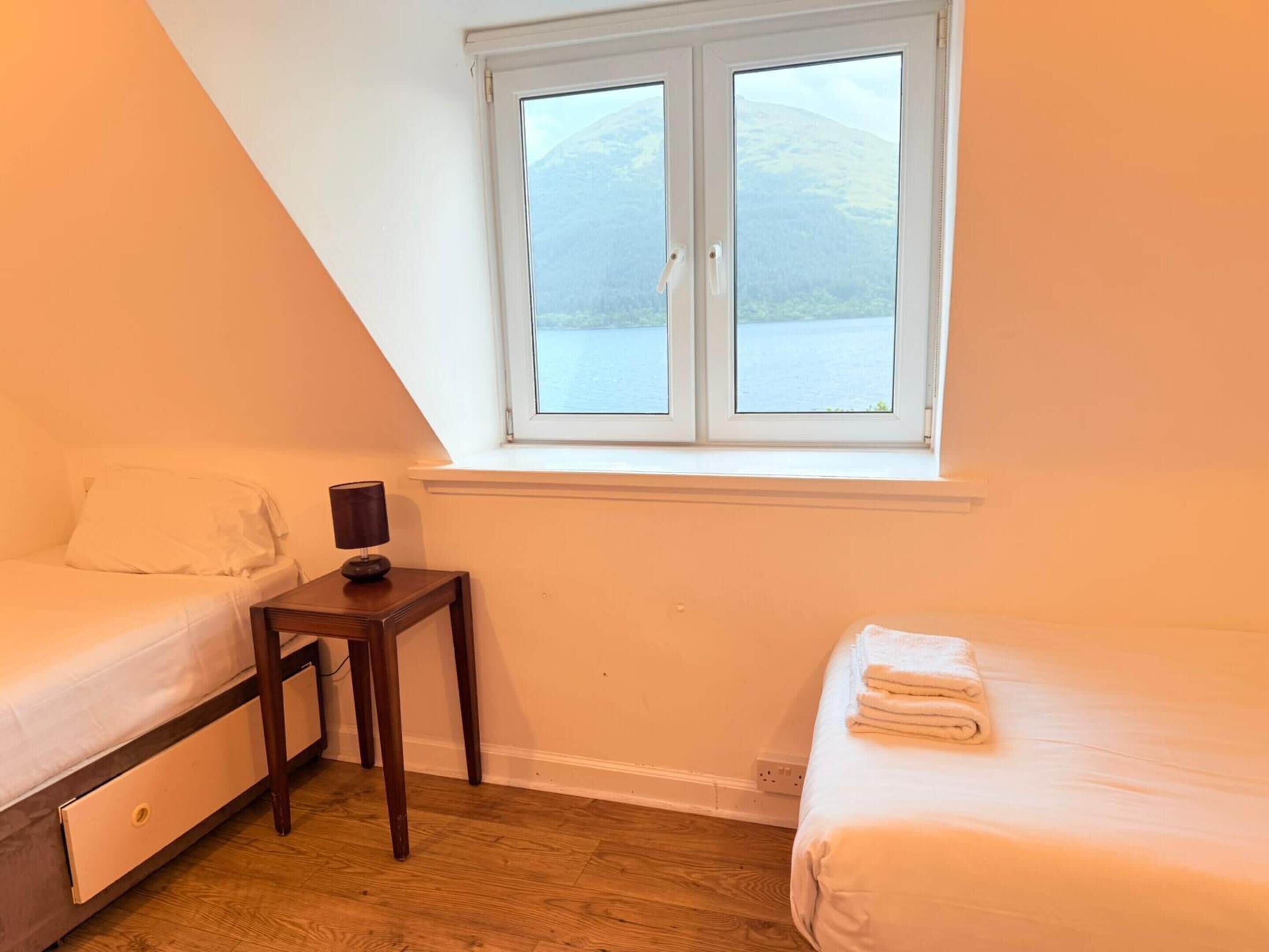 A small, well-lit room with two beds, a small table, and a window overlooking a body of water.