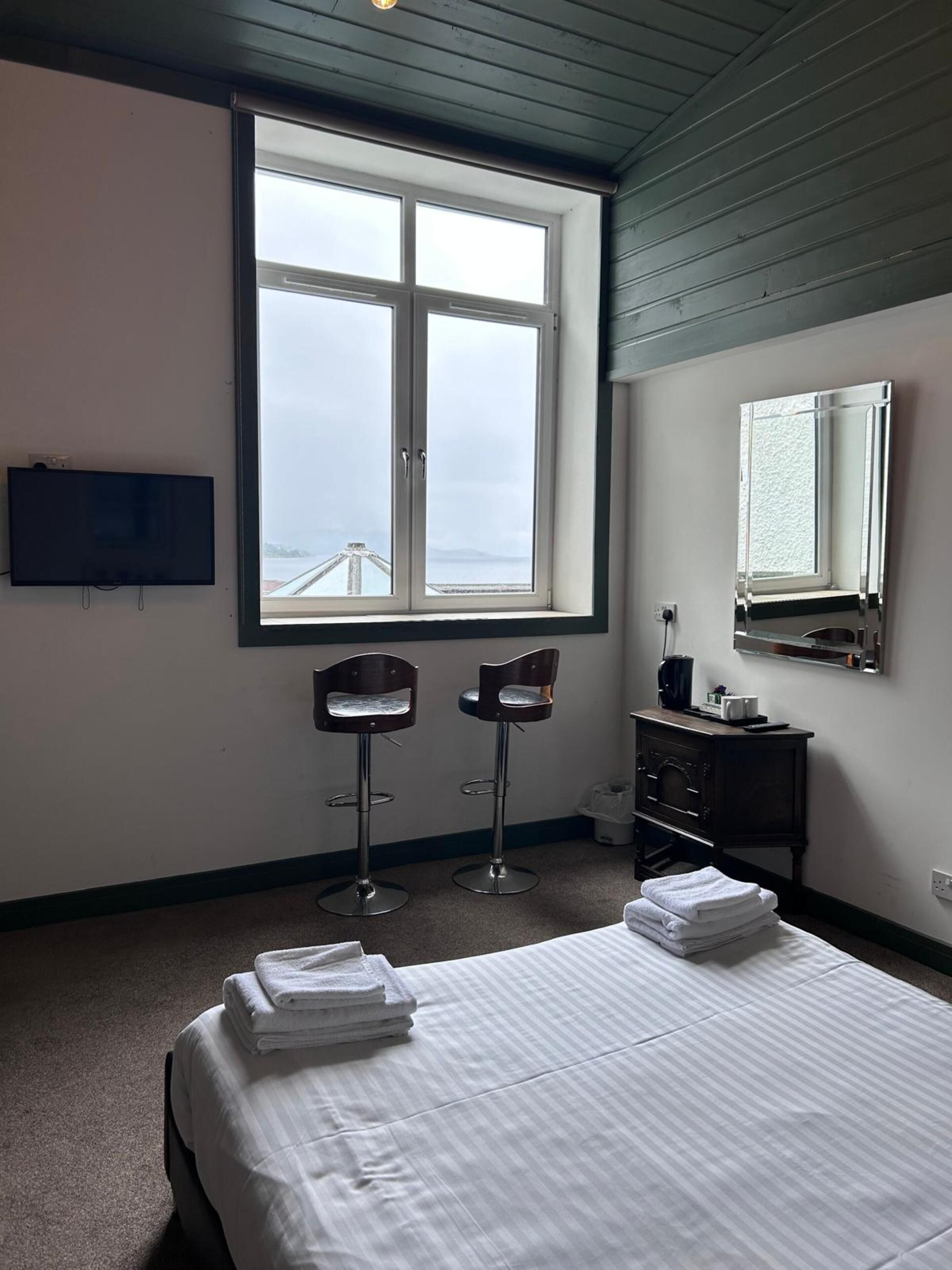 A hotel room with a bed, two bar stools, a small dresser, a TV, and a large window with a view of a bridge.