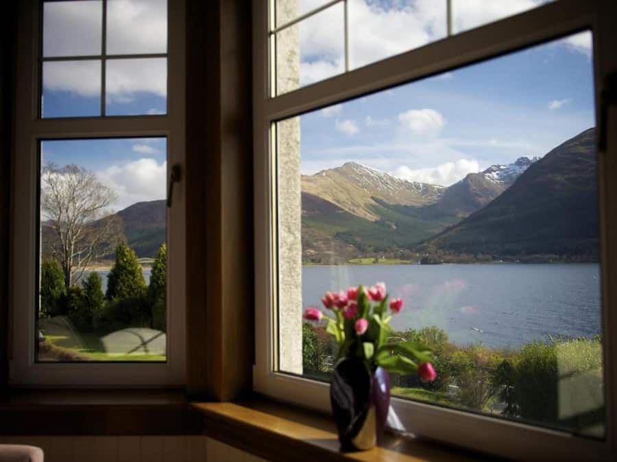 A window with a view of a lake and mountains, and a vase with flowers on the windowsill.