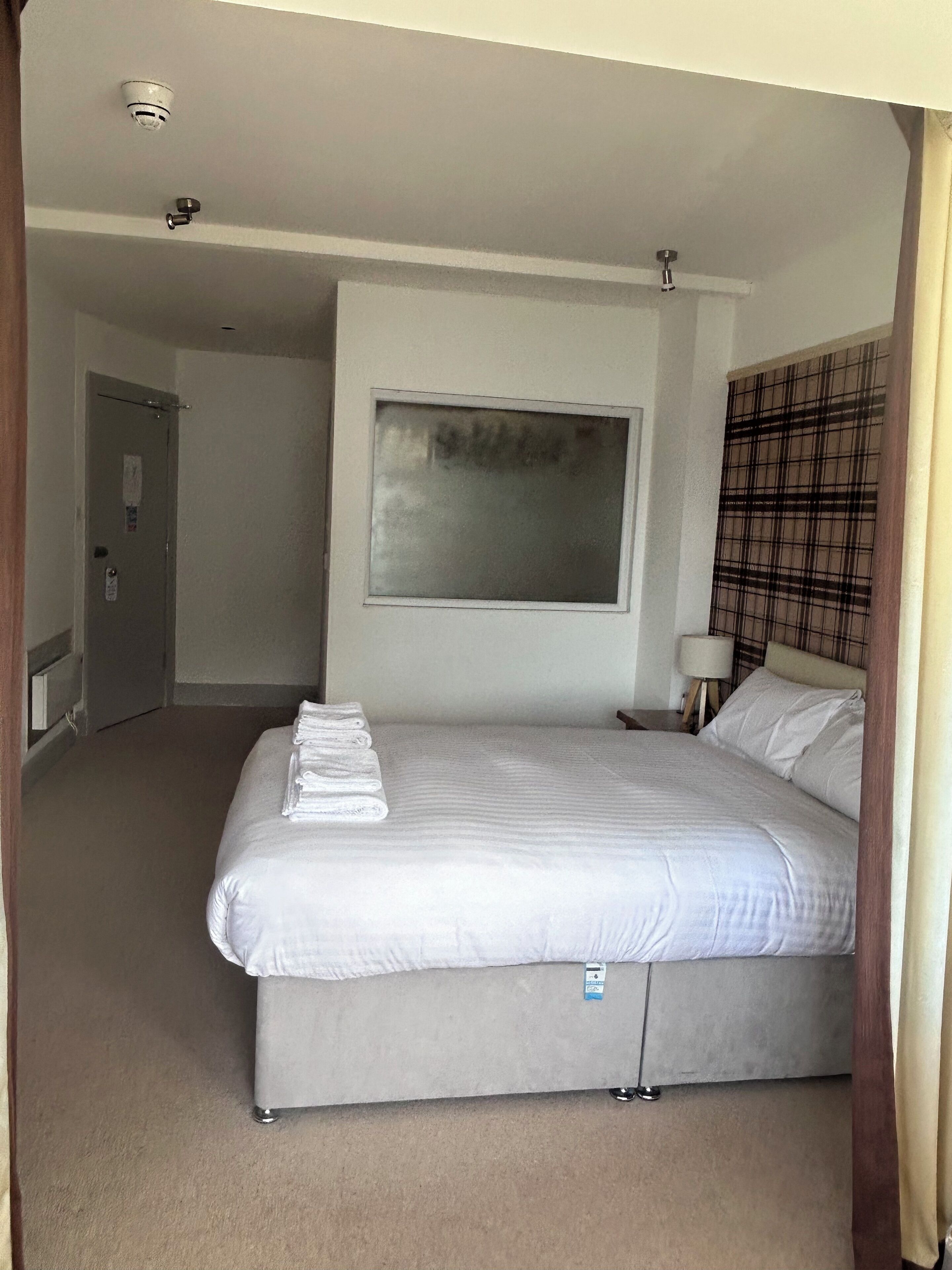 A hotel room with a large bed, a window, a door, and a wall-mounted light fixture.