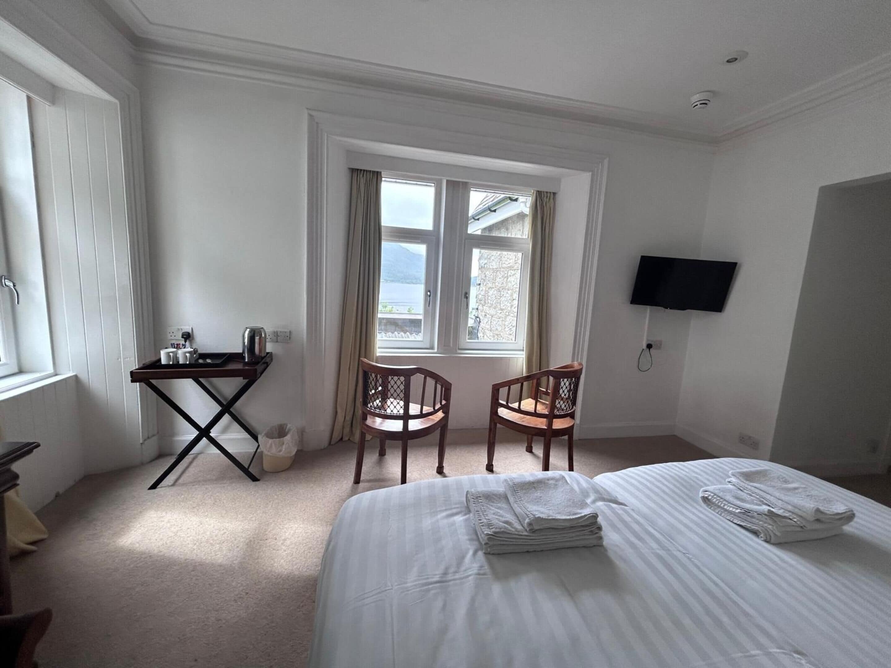 A hotel room with a bed, two chairs, a desk with a kettle, a TV, and a window with curtains.